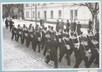 Uniformsklädda flottister marscherar längs med den snötäckta Drottninggatan. Gatan kantas av Sparres kanslibyggnad. Utanför byggnaden står militärer av högre grad.