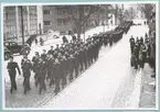 Ett långt paradtåg med uniformerade flottister som sträcker sig längs Drottninggatan nästan hela vägen bort mot Vallgatan. Kullerstenarna på gatan är täckta med ett tunt lager snö och vid sidan av, uppe på appareljen mittemot Bataljon Sparre, står civila åskådare.