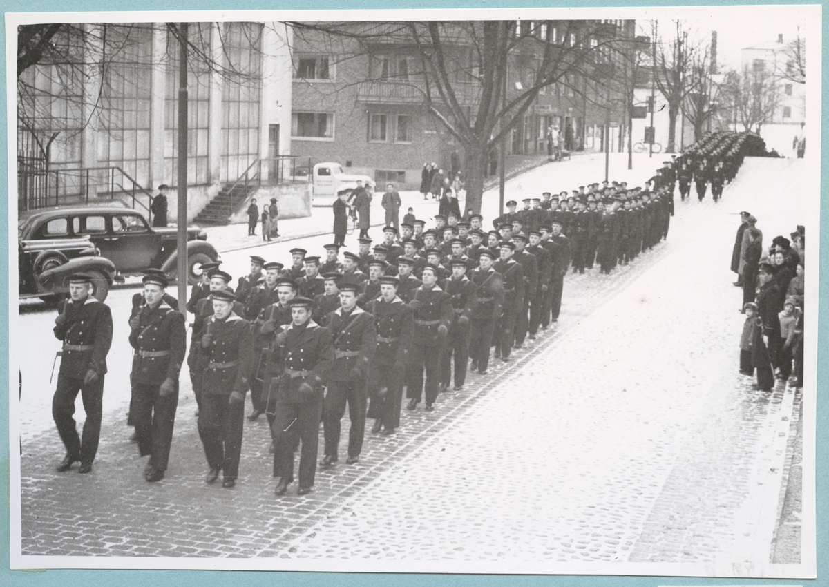 Uniformsklädda flottister marscherar längs med den snötäckta Drottninggatan. På gatan står både militära och civila åskådare. På gatan syns även enstaka motorfordon. Gatan kantas av lägenhetshus samt simhallen tillhörande bataljon Sparre.