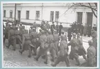 Uniformerade soldater ur armén marscherar längs Drottninggatan. Kullerstenarna på gatan är täckta av ett tunt lager snö. De marscherar förbi Bataljon Sparre, och utanför bataljonens portar står en grupp högre officerare givakt.