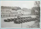 Flera militärlastbilar står parkerade på bataljon Sparres kaserngård. Kaserngården avgränsas i söder av simhallen och exercishuset tillhörande bataljonen. På gården står även flera militärer.