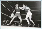 Två boxare i full färd med en boxningsmatch. Den ena mannen håller på att dela ut ett slag. De befinner sig i en boxningsring och utanför den skymtar åskådare.