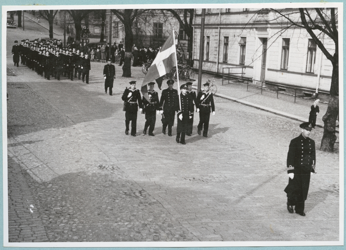 Uniformsklädda flottister marscherar längs Drottninggatan under ledning av en kapten och en löjtnant med en flagga. Den stenlagda gatan kantas av bataljon Sparres kanslibyggnad. 
Kyrkparad för nyinryckta värnpliktiga.