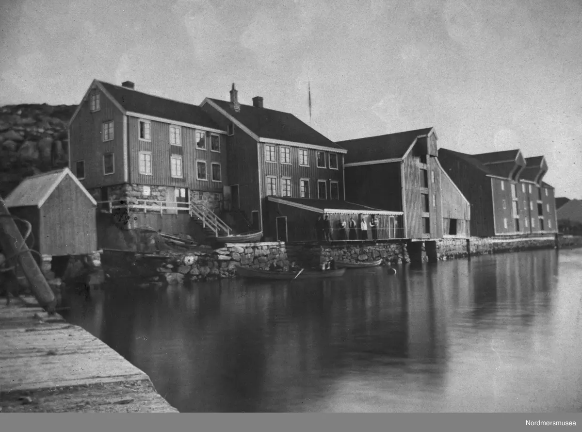 Fra venstre: skipper Strands vedbu, Strands bolig, bøkker Eriksens bolig, bøkkerverksted med skur foran, Eriksens brygge med tønne-stavskjul, Hjelkrembrygga (inntil 1904 Christie-brygga). På kaia og i båten sannsynligvis medlemmer av familiene Strand og Christoffersen. Matr.nr. 482a står skrevet med blekk på papplaten over bildet. En pil er tegnet på selve bildet og peker ned på Strandhuset. Det miderste bolighuset og bøkkerbrygga kalles idag, 2008, for Woldbrygga. Brygga ytterst til høyre kalles fortsatt for Hjelkrembrygga og eier er Kristiansund kommune. De andre eiendommene eies av Nordmøre Museum. Fra Nordmøre Museums fotosamlinger.