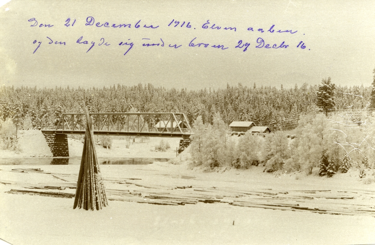 Plassen bru. Tømmer i forgrunnen. Hus i bakgrunnen. Vinter. Elva åpen under brua.