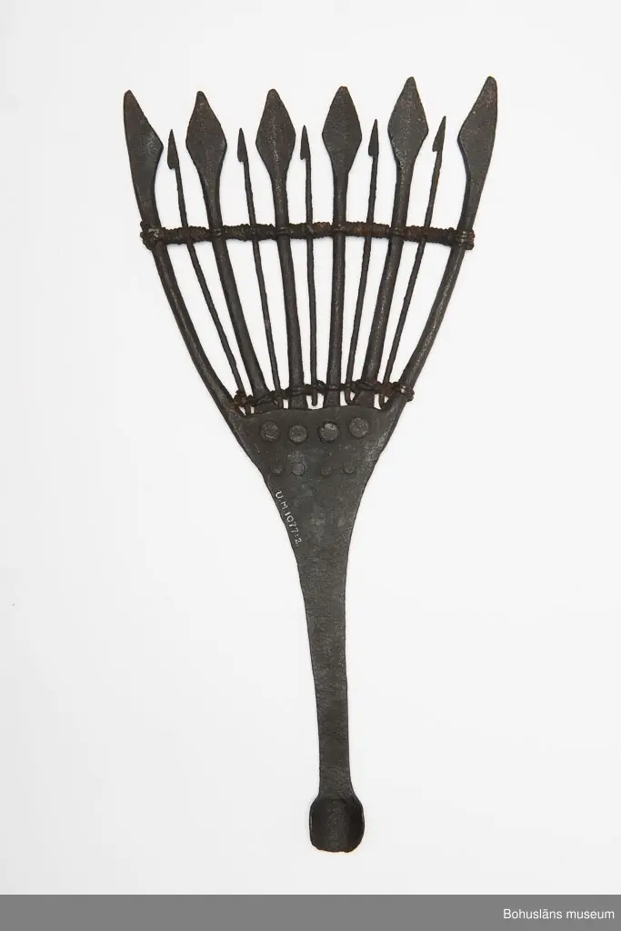 Gaffelliknande. Elva tenar, varav sex är tjocka och avslutas med ett blad. Fem stycken är betydligt tunnare, lite kortare och hullingförsedda. De tjocka och de tunna tenarna är placerade varannan bredvid varandra. Tenarna löper ut från ett mittparti. De fyra inre tjocka
tenarna är fastnitade. De två yttre tjocka tenarna är smidda ur skaftänden. De tunna tenarna sitter fastsurrade med snöre både upp och nertill. Skålform i skaftänden.
Skaft saknas.
Detta är ett oäkta grenljuster enligt Håkan Fernholms indelning av ljuster, tidskriften Folk-Liv 1942.
Ljustret har en ålderdomlig karaktär och kan vara äldre än 1700-tal,
Se även Haneson & Rencke, Bohusfisket, Göteborg 1923, sidan 228, 231 - 234. Föremålet är avbildat och onmämnt i en artikel om ljuster av Börje Rosén i Bohusläns Hembygdsförbunds Årsskrift 1961.
Givaren handlade med skrot.
Inventerat 1996-08-12 GH.

Ur handskrivna katalogen 1957-1958:
Nitton juster se vidare sid 485
Ljuster av järn, fr. Bohuslän
L.43,5 Br. 21 cm; 11 taggar varannan m. hullingar, varannan m. flata, trubbiga spetsar; virat m. snöre. Helt.

Lappkatalog: 48