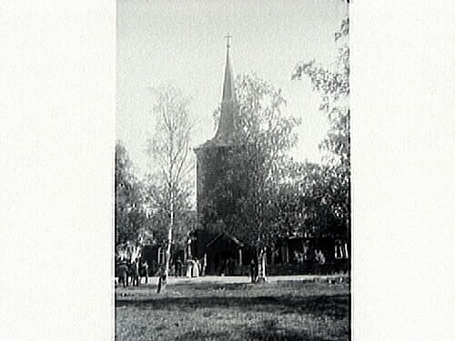 Nysunds kyrka i Närke med björkar i förgrunden. Utanför kyrkmuren är människor på väg in till kyrkan.