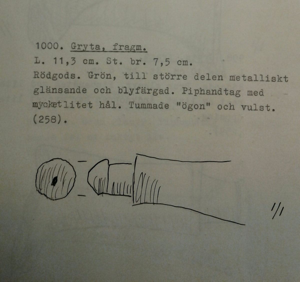 Avsmalnande piphandtag samt delar av kärlets liv i yngre rödgods. Sannolikt till en trefotsgryta. Handtaget är avsmalnande med en utsträckt knopp och ett mycket litet hål i änden. Typ D2, år 1425 - 1550. Vid övergången till kärlets buk är tummade "ögon" och en smal vulst. Insidan av buken är grönbrun, till större delen metalliskt glänsande. Tillverkad i södra Skandinavien.