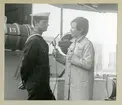 Bilden föreställer en kvinna med en mikrofon som intervjuar kronprinsen Carl Gustaf, som står klädd i uniform. Bilden är tagen under minfartyget Älvsnabbens långresa 1966-1967.