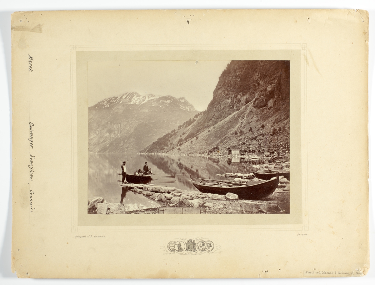 NR. 374 (13,5X18) 
Merok, Geiranger. Et følge av mennesker som er ute i robåt på fjorden.
bebyggelse og båter i strandlinjen, det hele er omkranset av fjell.