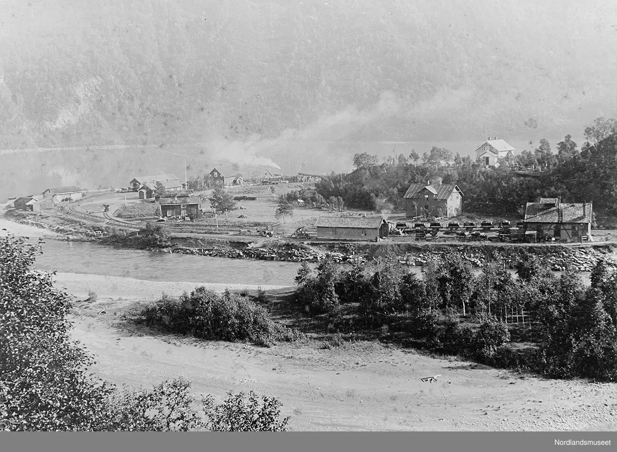 Bebyggelse ved et vatn med jernbane. Sulitjelmabanen, Sjønstå stasjon ved Øvrevatnet. Skogkledd fjellside i bakgrunnen. I forgrunnen noe av vatnet. Midt på bildet bebyggelsen og jernbanen m/stasjonen. Et malmtog på stasjonen til høyre.
