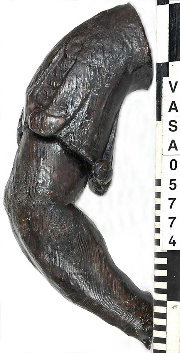 Del av skulptur. Arm till Stora krigaren på babord sida. Mycket kraftig. Bruten. Saknar hand samt en del av klädnadens ärm. Vackert snidad. Konstnären har använt sig av träets ådring för att få fram musklernas form.

Text in English: Arm belonging to sculpture. Very worn, broken.