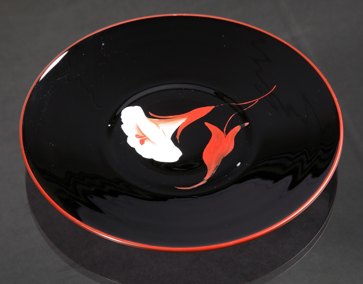 Assiett.
Teknik: Pressat, slipad bottenring, målad.
Färg/form: Svart degelglas, rund. Målad i rött, vitt och gult.
Dekor: Blommor likt blomman för dagen.
Målad av Karl Diessner (målning identifierad av Susann Diessner 2021-04-20).
Givarens mormor berättade att hennes morbror, Hans Teodorsson eller hans son George Hansson tillverkade föremålen. De arbetade på Eda innan de flyttade till Småland.
Givarens (Sivs) mormor fick föremålen av Hans eller George.