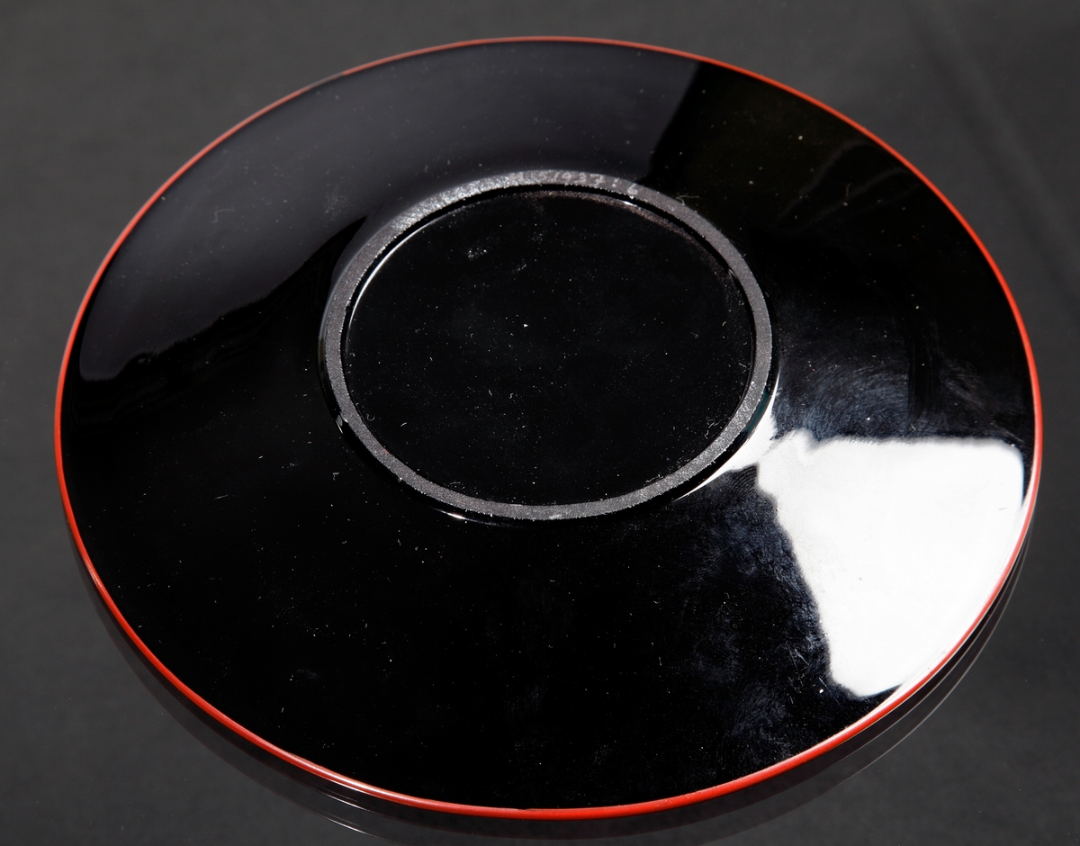 Assiett.
Teknik: Pressat, slipad bottenring, målad.
Färg/form: Svart degelglas, rund. Målad i rött, vitt och gult.
Dekor: Blommor likt blomman för dagen.
Målad av Karl Diessner (målning identifierad av Susann Diessner 2021-04-20).
Givarens mormor berättade att hennes morbror, Hans Teodorsson eller hans son George Hansson tillverkade föremålen. De arbetade på Eda innan de flyttade till Småland.
Givarens (Sivs) mormor fick föremålen av Hans eller George.