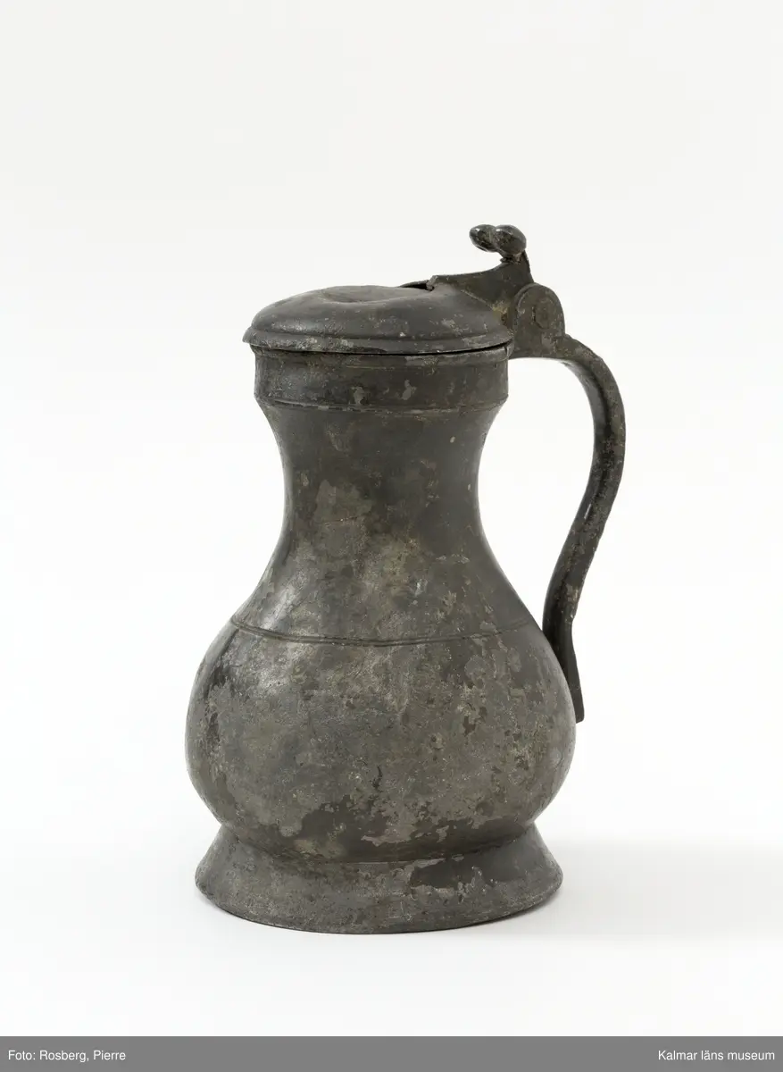 KLM 1886. Kanna, hankkärl. Av tenn. Hankkärl, bukig form, försedd med lock och handtag men utan näbb. Grepen ornerad på utsidan med bladranka som upptill avslutas med en djurliknande figur. Bottenmedalj i kannan, i form av korsfästelsemedalj. Spår efter förlorad lockmedalj, på lockets insida.