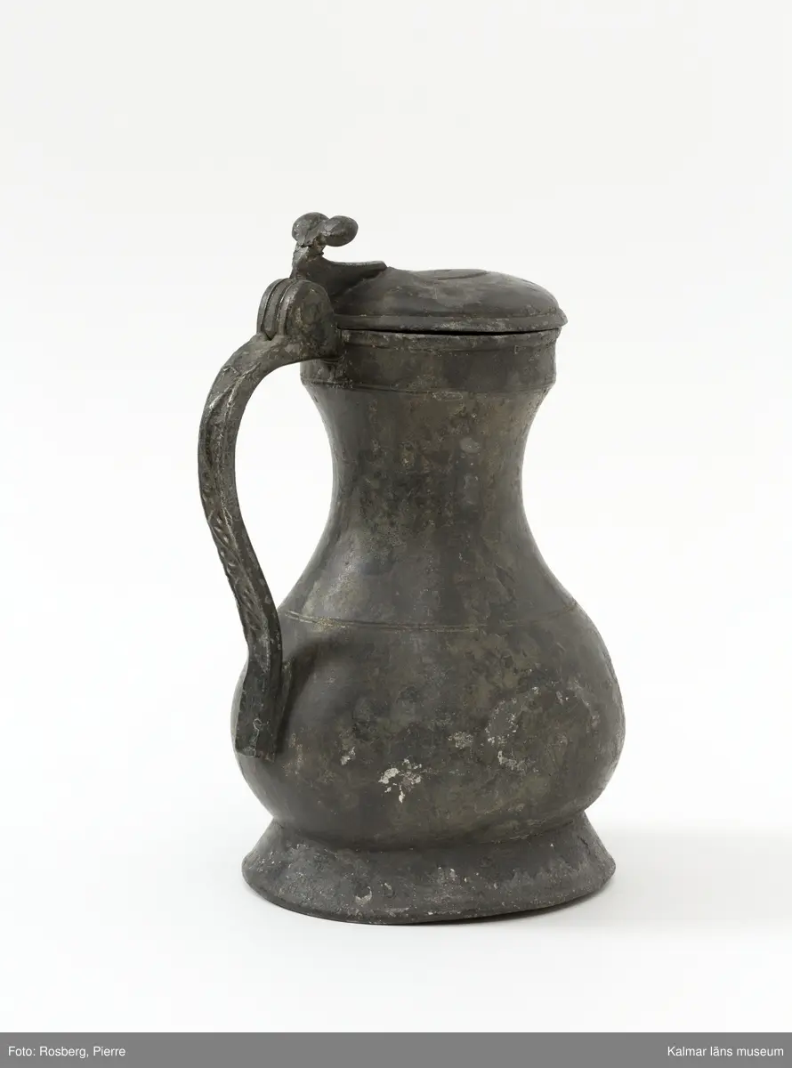 KLM 1886. Kanna, hankkärl. Av tenn. Hankkärl, bukig form, försedd med lock och handtag men utan näbb. Grepen ornerad på utsidan med bladranka som upptill avslutas med en djurliknande figur. Bottenmedalj i kannan, i form av korsfästelsemedalj. Spår efter förlorad lockmedalj, på lockets insida.