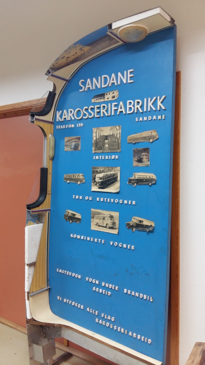 Reklameinstallasjon. Visningsmodell. Finèrplate påmontert snitt av karosseri på buss, med isoporisolasjon og gummlistar rundt vindauge, utside med pyntelistar, innside med kupèlys og vindauge med vindaugsisolasjon. Snittet går i ei boge på eine langsida og opp rundt langs øvste kanten, og er festa til ei finèrplate. Plata er blå på framsida, med pålimte bilete av bussar og andre bilar og tekst i gipsbokstavar. Installasjoen står på eit bein og ei støtte av metall.

 karosseriet på ein buss