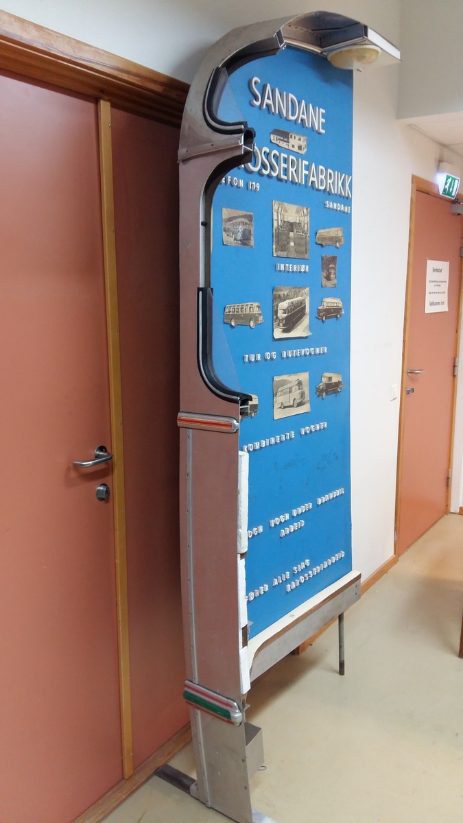 Reklameinstallasjon. Visningsmodell. Finèrplate påmontert snitt av karosseri på buss, med isoporisolasjon og gummlistar rundt vindauge, utside med pyntelistar, innside med kupèlys og vindauge med vindaugsisolasjon. Snittet går i ei boge på eine langsida og opp rundt langs øvste kanten, og er festa til ei finèrplate. Plata er blå på framsida, med pålimte bilete av bussar og andre bilar og tekst i gipsbokstavar. Installasjoen står på eit bein og ei støtte av metall.

 karosseriet på ein buss