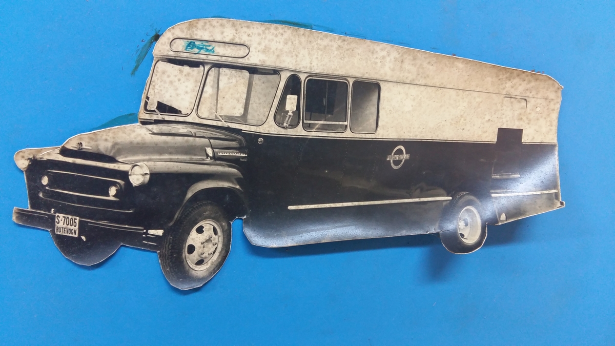 Reklameinstallasjon. Visningsmodell. Finèrplate påmontert snitt av karosseri på buss, med isoporisolasjon og gummlistar rundt vindauge, utside med pyntelistar, innside med kupèlys og vindauge med vindaugsisolasjon. Snittet går i ei boge på eine langsida og opp rundt langs øvste kanten, og er festa til ei finèrplate. Plata er blå på framsida, med pålimte bilete av bussar og andre bilar og tekst i gipsbokstavar. Installasjoen står på eit bein og ei støtte av metall.

 karosseriet på ein buss