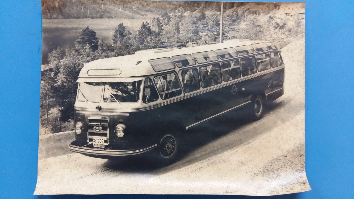 Reklameinstallasjon. Visningsmodell. Finèrplate påmontert snitt av karosseri på buss, med isoporisolasjon og gummlistar rundt vindauge, utside med pyntelistar, innside med kupèlys og vindauge med vindaugsisolasjon. Snittet går i ei boge på eine langsida og opp rundt langs øvste kanten, og er festa til ei finèrplate. Plata er blå på framsida, med pålimte bilete av bussar og andre bilar og tekst i gipsbokstavar. Installasjoen står på eit bein og ei støtte av metall.

 karosseriet på ein buss