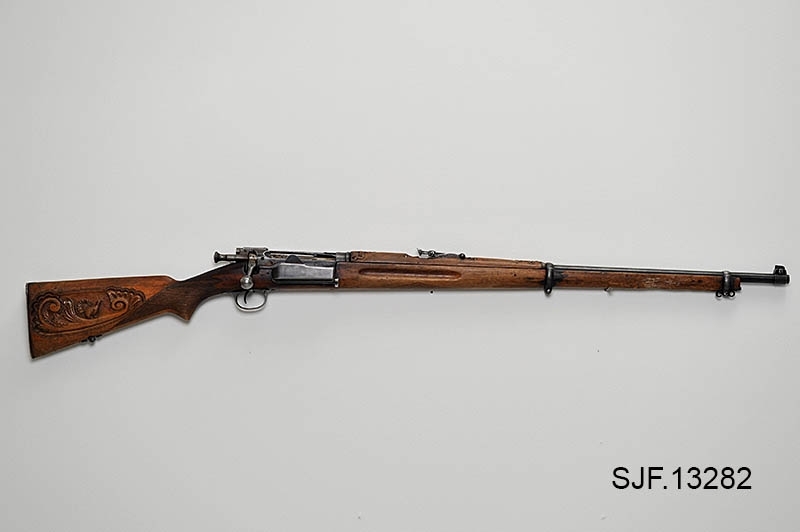 Krag-Jørgensen rifle med kaliber 6, 5 x 55. Geværet har pistolgrep. Magasinkapasitet på 5 patroner. Kolben er brunbeiset / oljet. Skjeftet er dekorert med forseggjorte utskjæringer.  Kolbekappen av stål har innfelt et lokk. Om man åpner lokkket kommer man til et "oppbevaringsrom" i kolben. Treverket som omslutter undersiden av geværløpet har noen sprekkdannelser.  Geværet er utstyrt med høydejusterbart skur og sidejusterbart korn. 