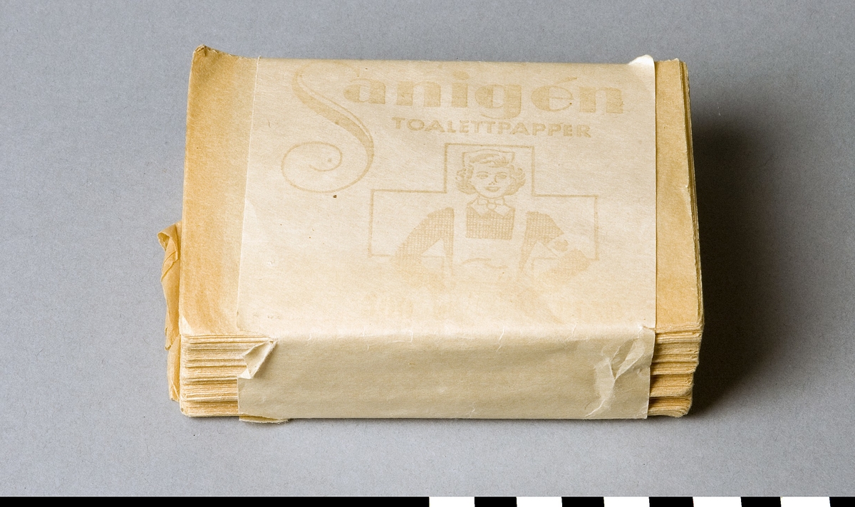 Förpackning med toalettpapper i bladform. Vit pappersremsa omslutande toaletpappersark. Text: Sanigen TOALETTPAPPER + bild av sjuksköterska framför ett kors. 200 blad.
