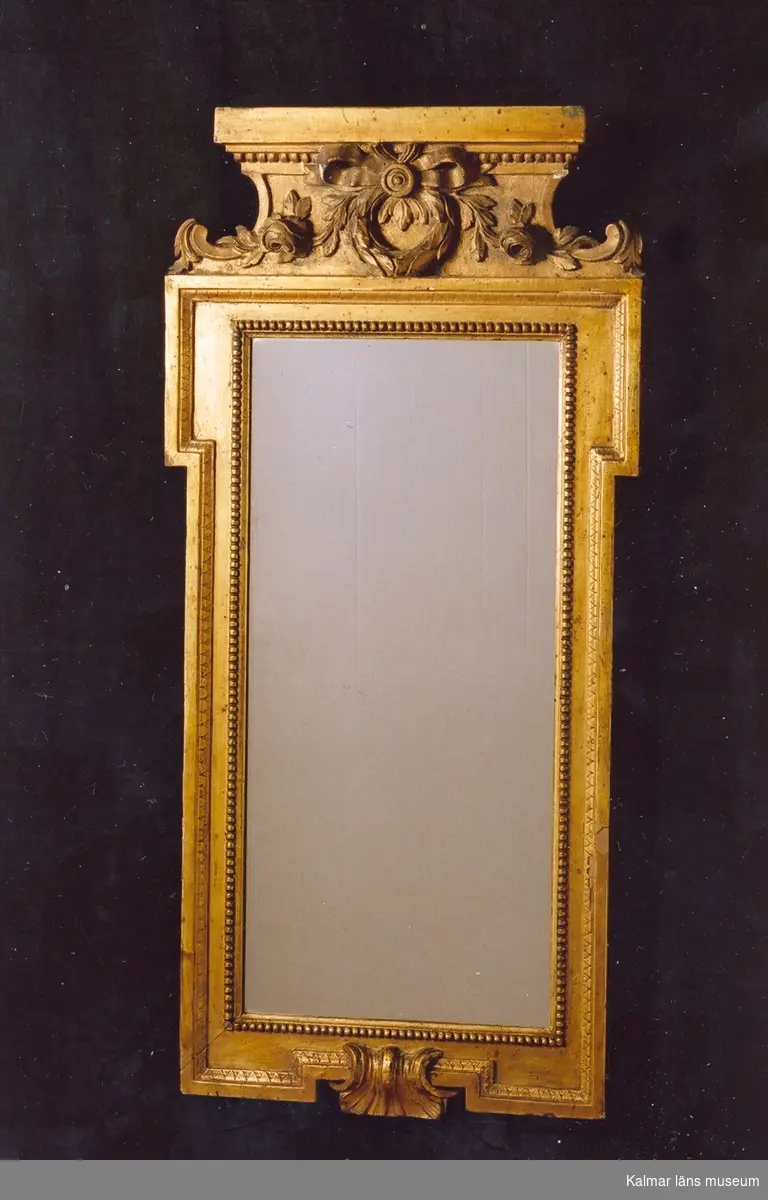 KLM 31938. Spegel. Förgylld bred ram av trä med pärlstav närmast spegeln och ytterkanten med äggstavsmönster. Krönet försett med en ros på vardera sida om en lagerkrans bunden med en rosett. Allt i relief. Svårläsbar stämpel på ramens baksida tockh..hal, 780, kan tolkas som: Stockholm Hallrättsstämpel, 1780. Nyare spegelglas.