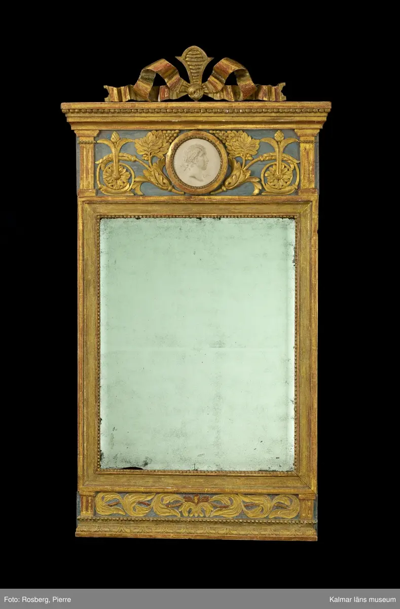 KLM 45592. Spegel, väggspegel. Utförd i sengustaviansk stil. Skuren ornamentik i trä och i gjutet pastellage, förgylld med slagmetall. Ovanför glaset en rund medaljong med relief. Fasetterat originalglas. Träskuret krön i form av en avtagbar rosett.
