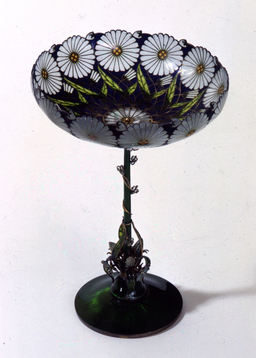 Skålformet vase med lang, tynn stett og opphøyet fotstykke. Foten er laget i sølv og grønn emalje. Fra denne slynger det seg blomster, knopper og bladverk oppover stetten. Oppunder den flate glasskålen er det tre blomster, knopper og blad. I selve skålen er det innerst en rosett med gullkule i midten. Videre er det samme mønsteret gjentatt på mørk blå bunn; to grønne blad, en knopp og øverst en utsprungen margeritt.