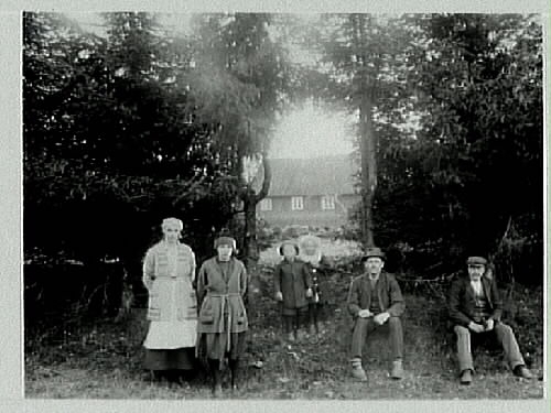 En grupp människor vid en hög granhäck. Genom en öppning i häcken syns en lantgård i bakgrunden. Två kvinnor, två män och två barn.