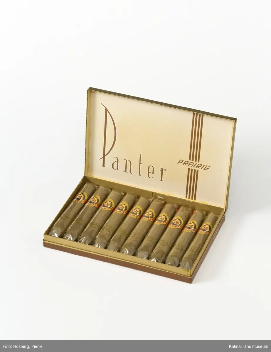 KLM 45058:38. Cigarrask, av papp och tobak. Cigarrask i brunt, guld och vitt. En panter som ligger och spanar på locket och text: Panter prairie. Förpackningen innehåller 10 inplastade cigarrer med gördlar med en bild på ett panterhuvud och texten panter. Bruten förpackning men med alla cigarrer kvar i förpackningen.