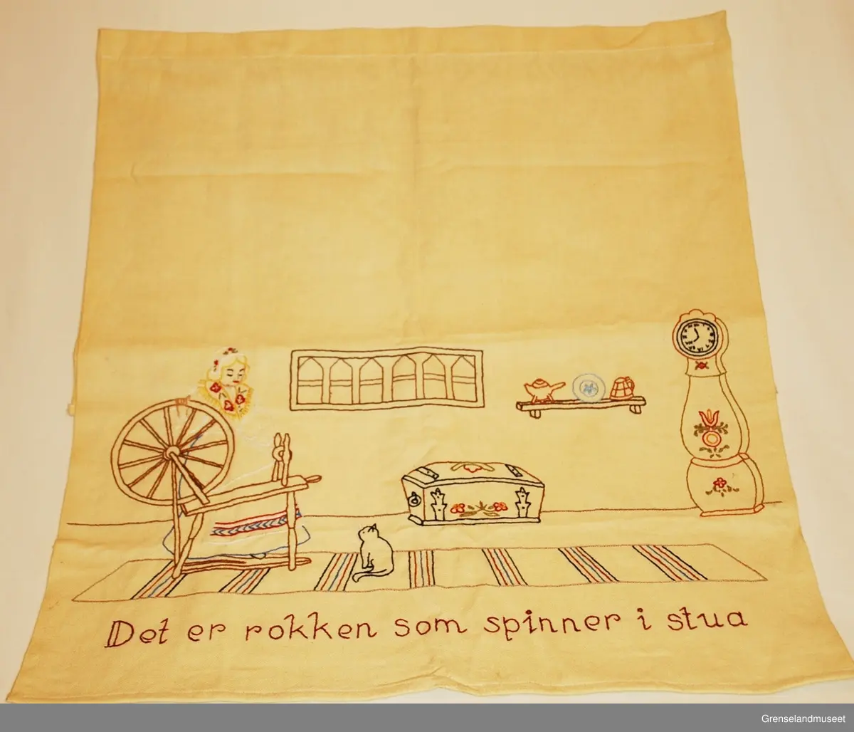 Brodert på gult bomullsstoff. Interiør med kvinne i bunadlignende drakt som spinner på rokk, kiste, gulvur, fillerrye og katt. Kontursting og tubgesting. "Det er rokken som spinner i stua"