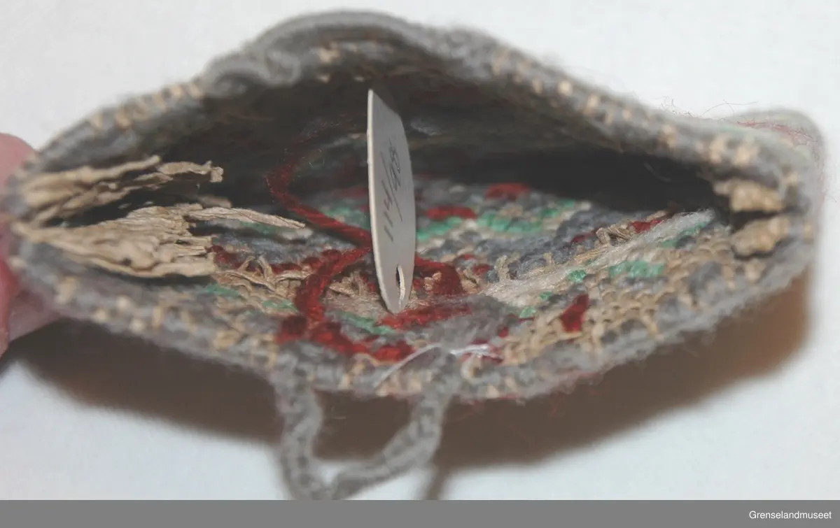 Brodert på stramei. Grå med hvite og rød sikksakkbord.
På den ene siden brodert: ERNA og 1944. På den andre siden: LUBECK
