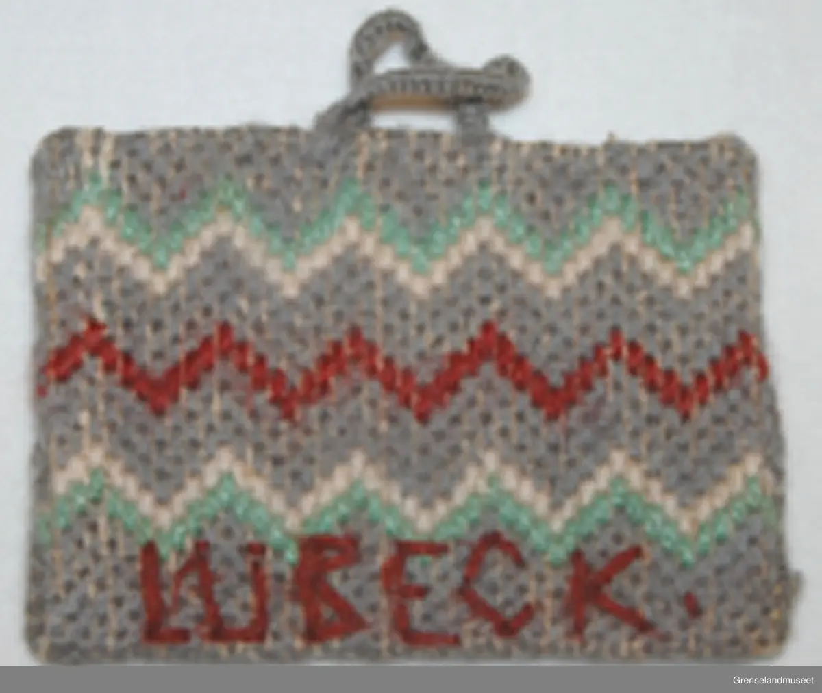 Brodert på stramei. Grå med hvite og rød sikksakkbord.
På den ene siden brodert: ERNA og 1944. På den andre siden: LUBECK