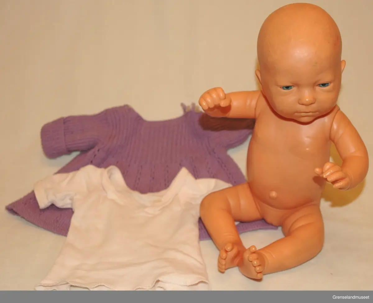 Plastik/gummi med bevegelige armer, hode og ben. Påkledd med babyklær: trøye og lilla strikket kjole.