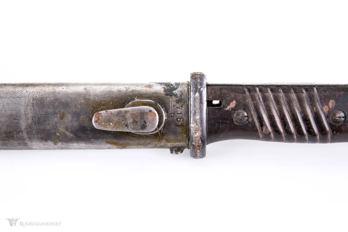 Metallhåndtak med lang pommel og geværfeste med fjæret knapp i bakkant. Serratert tregrep som er festet med 2 skruer. Langt spisst stålblad med egg og blodrand. Metallslire med kule enden og krok for festing til grunnutrustning/belte.