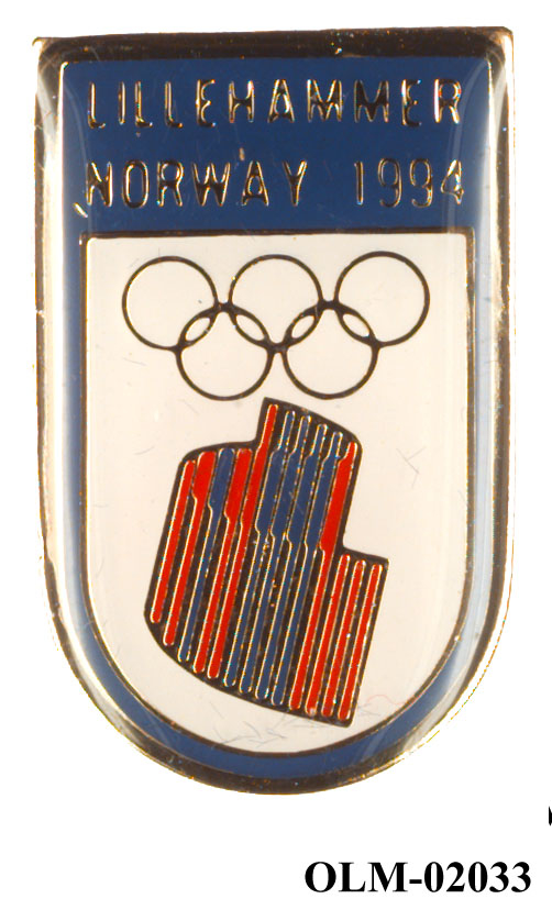 Skjoldformet merke med de olympiske ringer og nordlyslogo som vender nedover på hvit bakgrunn. Tekst øverst i sølvbokstaver på mørkeblå bakgrunn.