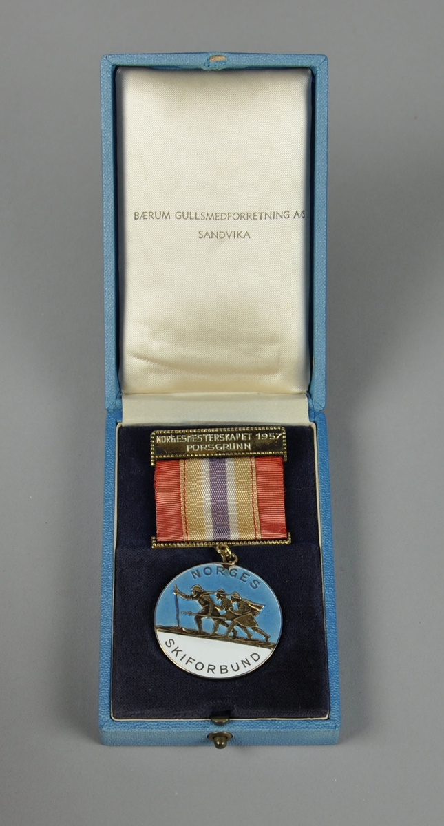 Medalje i etui.  Motivet viser tre skiløpere (Arnljot Gelline, Torodd Snorreson og følgesvenn), på blå og hvit bakgrunn. Bånd i rødt, hvitt og blått.