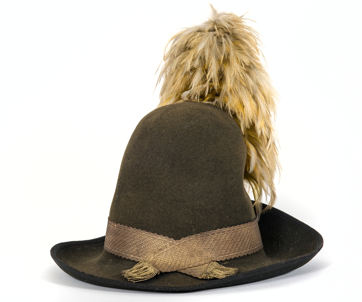 Hatt av svart kläde med hög kulle, breda brätten. Uppvikt framtill för fäste av gul sidenkokard och plym. Ofodrad. Svettrem av brunt läder. Bred guldgalon  med fransade  ändar runt hatten. Plym av gul hönsfjäder.