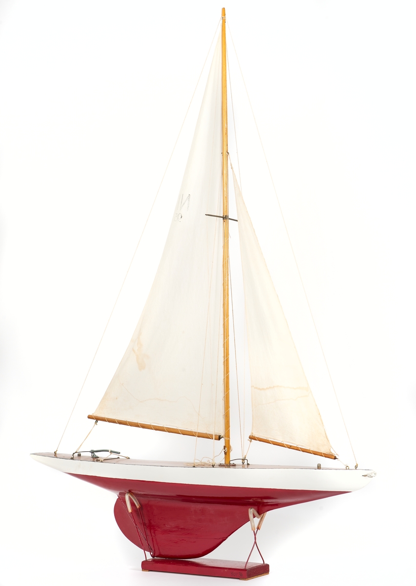 Modell av segelbåt, leksak. Tillverkad av NK senast 1935. 
Vitt skrov med röd bottenmålning. Fernissat nåtat däck och blyköl.