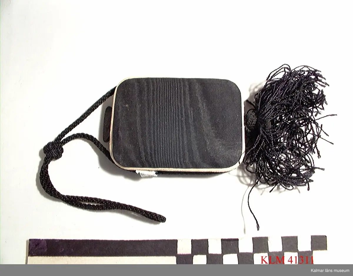 KLM 41311 Puderdosa, textil, spegelglas. Rektangulär, klädd med svart tyg, kantad med svart snodd, tofs. Inuti två runda fördjupningar med vitt respektive rött puder/rouge, rund vippa av tyg, spegel i locket.