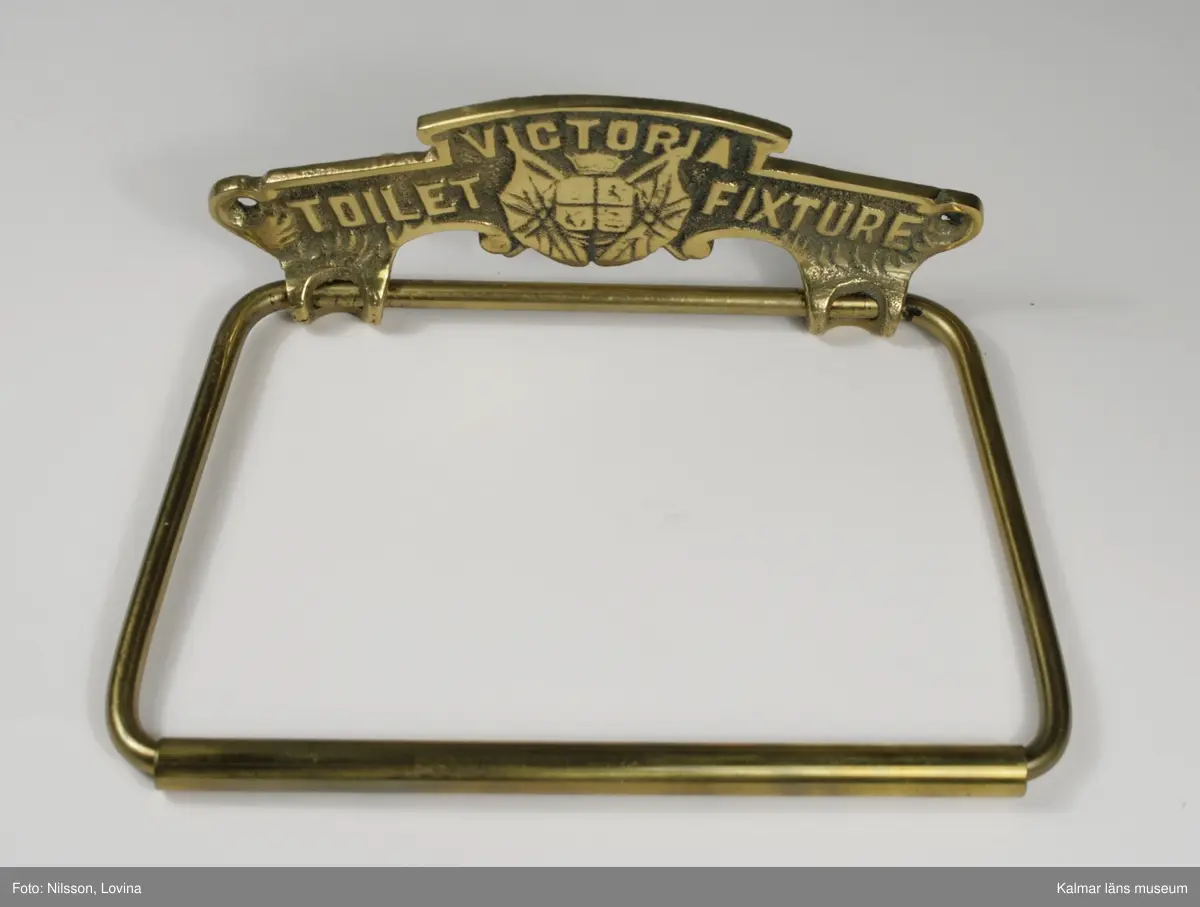 KLM 44292:3. Toalettpappershållare av mässing. Gjuten. Tillverkad av Lindstedt & Son, Kalmar, tillverkaretikett på baksidan. Gjuten text på hållarens framsida: Victoria, Toilet, Fixture.
