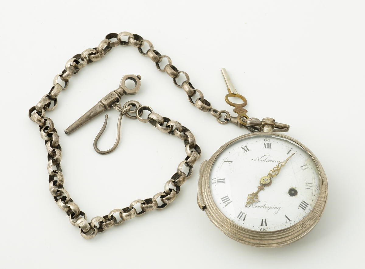 Fickur i silver.
Boett i silver, förgylld invändigt med text: "Eric Nohrman, Norrköping, No 381". Vit emaljerad urtavla med romerska siffror och förgyllda, genombrutna visare. Text; "Nohrman, Norrköping". 
Tillhörande kedja, se M 66156-2.
Inskriven i huvudkatalog 2017.