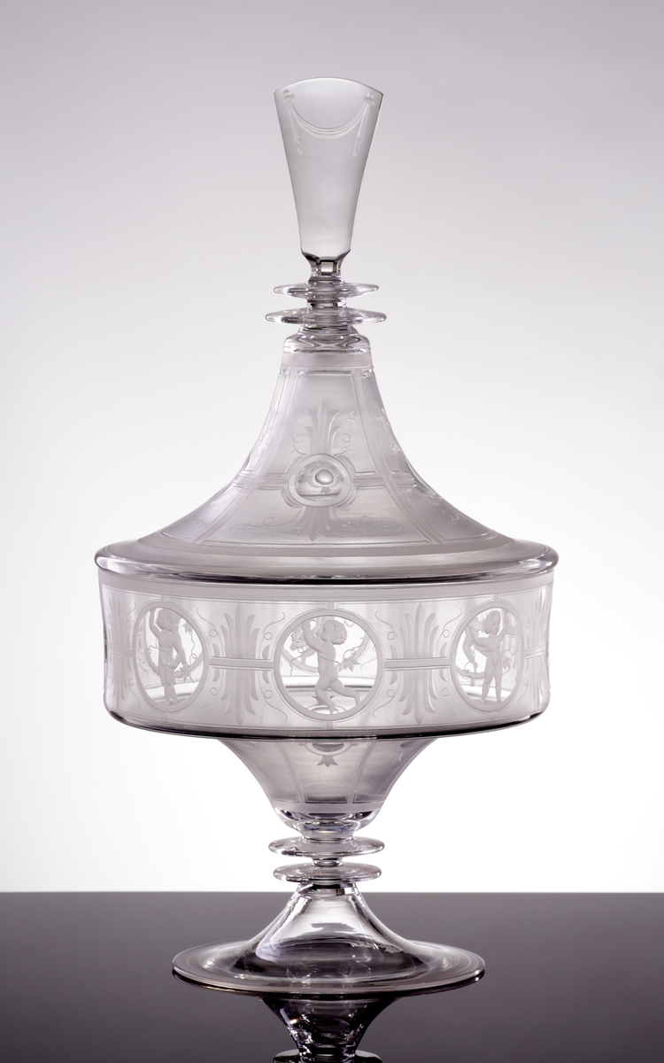 Skål eller urna med lock.
Skålen har en låg cylindrisk form som neråt smalnar av till en strut. Benet består av två ringar, och foten är trattformad. Locket är trattformat, där knoppen inleds med två ringar och slutligen en konisk knopp.
Skål och lock är mattsyrade i olika geometriska fält, vilka sedan graverats med organiska motiv. Skålen har runda fält vari åtta olika puttimotiv med bacchusinspiration graverats. På locket finns obearbetade "fönster", och på knoppen ett graverat draperi.