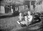Två flickor, den ena Marit Liljefors, klädda i folkdräkt, sitter på gräsmattan ute i trädgård, sannolikt Svensgården, Dalarna