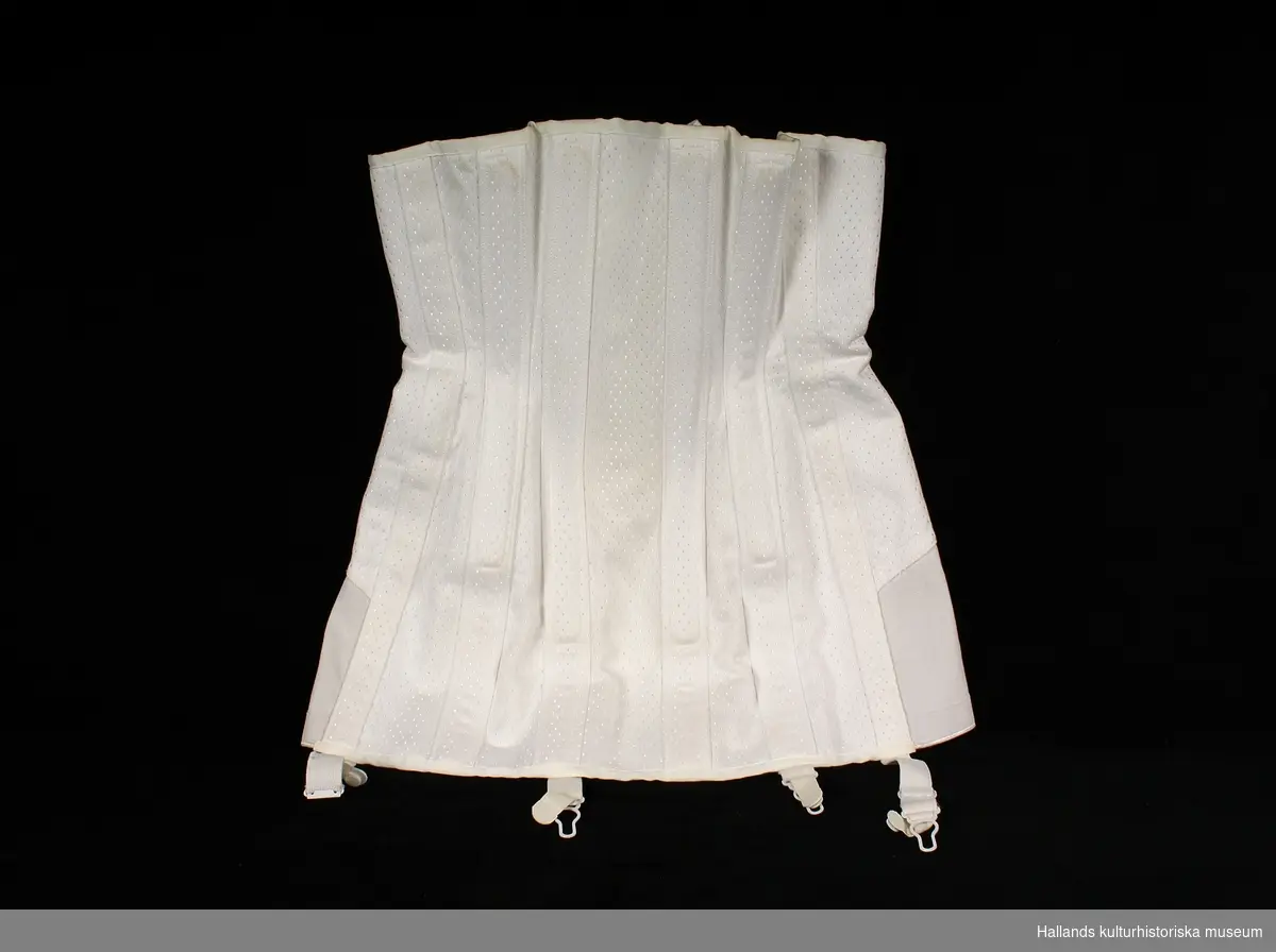 Korsett av vitt grövre atlassiden med vävt mönster. Isydda metallstöd för att ge korsetten stadga. Snörning i ryggen. Fyra strumpebandshållare.