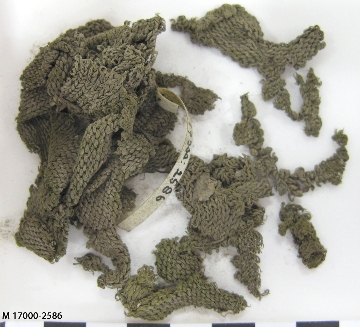 Flertalet mindre grågröna stickade fragment eventuellt av siden, stickade i slätstickning. Tråd samt mask och varvtäthet: 2-trådigt Z-spunnet, S-tvinnat masktäthet: 4-6 maskor/cm varvtäthet: 5-7 varv/cm.