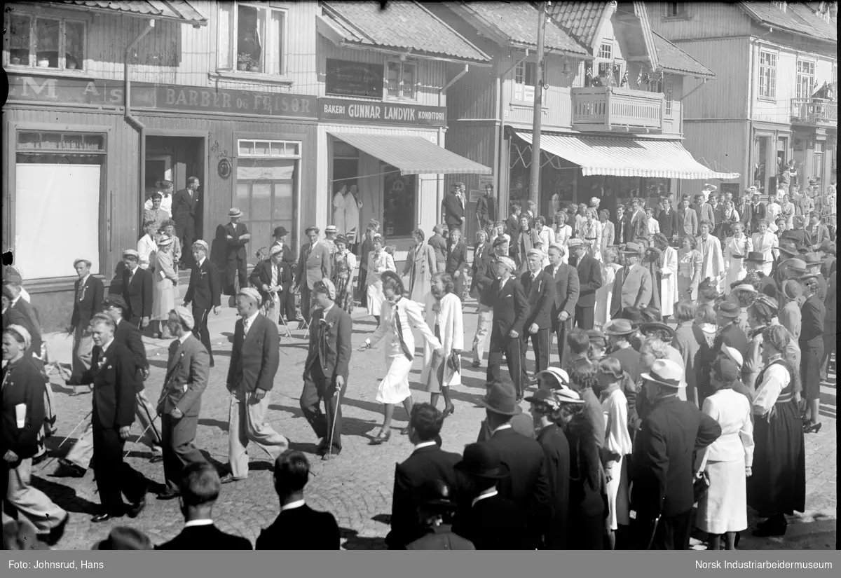 17. mai feiring i Notodden sentrum. Folketog med blant annet russ gående gjennom byens gater. Butikkvinduer for Barber og Frisør, Bakeri Gunnar Lindvik.