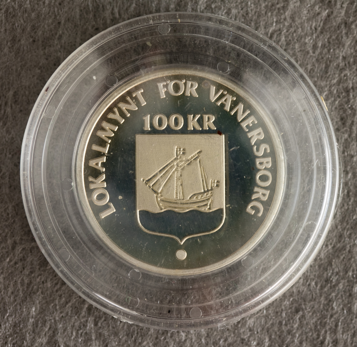 Ett lokalmynt för Vänersborg, i silver, 2,8 cm i diameter. På framsidan: text “Lokalmynt för Vänersborgs kommun 100  kr” samt en bild av en vapensköld med ett segelfartyg. 
På frånsidan: text “Sparbanken i Vänersborg 1822 - 1982” samt en bild av denna byggnad.
Myntet ligger i ett skyddsfodral.