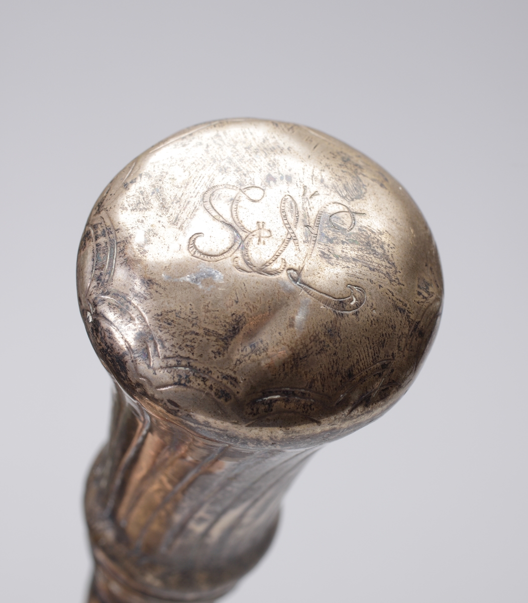 Promenadkäpp, s.k. spanskrör. 
Mörk, fernissad (?) käpp med delvis spiralvridet, snidat mönster och diamantskärningar. Knopp av silver med randad, något svängd dekor. På knoppens ovansida graverat ägarmonogram: "SEL" (?). Längst ner på käppen en skoning av ben. Stämplar längs nedre kanten av knoppen.